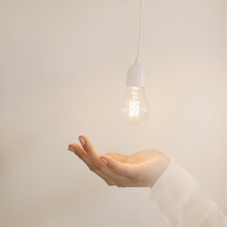 Une main ouverte sous une ampoule allumée suspendue, dans un décor minimaliste et lumineux, symbolisant une idée ou une révélation inspirante.